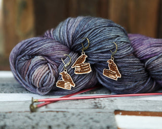 The Conspiracy of Ravens Knitting Progress Keepers, featuring three raven-on-books charms, is displayed with variegated purple and gray yarn and pink knitting needles on a rustic wooden surface.