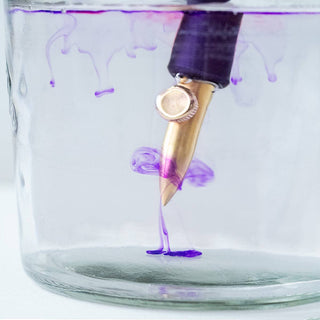 The One-Dip-Nib, featuring a gold calligraphy nib and purple handle, is dipped into a glass jar of water, where it releases vibrant purple ink that swirls and drifts through the clear liquid.