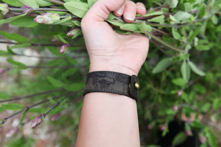 A hand holds leafy stems while wearing the Mushroom Leather Ruler Cuff, a leather woodland accessory featuring engraved animal designs and a brass button, set against a blurred background of green foliage and flower buds.