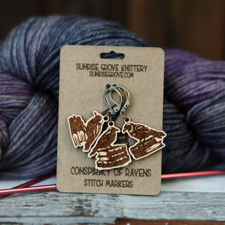 A set of maple wood Conspiracy of Ravens Knitting Progress Keepers with book designs, displayed on a Sunrise Grove Knittery card, shown with yarn and a needle—charming accessories for any knitting project.