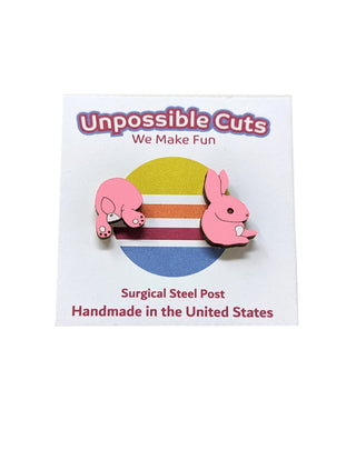 Two pink Bunny Butt Earrings, made with hypoallergenic surgical steel posts, are displayed on a colorful striped Unpossible Cuts card that reads “We Make Fun” and “handmade in the USA.”.