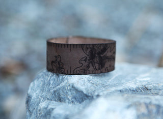 The Oak Leaves & Acorn Leather Ruler Cuff, featuring engraved ruler markings and intricate designs, rests on a gray stone surface against a blurred natural background.