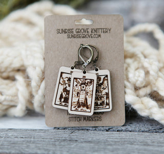 Three wooden Tarot Card Stitch Markers—Justice, Wheel of Fortune & The Chariot—each with a metal clasp, are displayed on a branded Sunrise Grove Knittery card. Unique crochet accessories for every maker; cream yarn blurred in the background.