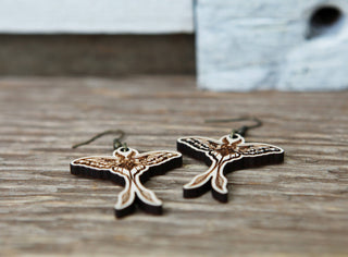 Luna Moth Maple Wood Earrings, featuring detailed wing engravings, are displayed on a rustic wooden surface.