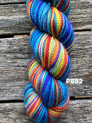 A skein from the KPM and KPPPM Variegated Color Collection in shade P882, featuring vibrant blue, red, orange, yellow, green, and purple tones, rests on a rustic wooden surface—perfect for hand knitting projects.