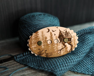 The Baba Yaga Row Counter, crafted from maple wood and featuring a cabin and trees design, rests on blue knitted fabric with a ball of blue yarn in the background.