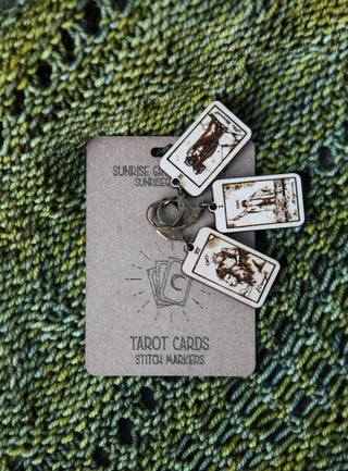 The 🔮 Tarot Card Stitch Markers – The Hermit, The Magician & Strength 🔮 set includes three illustrated markers for knitting, a progress keeper, and comes on a “Tarot Cards Stitch Markers” card displayed on green and blue knit fabric.
