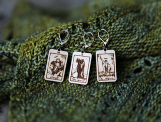 🔮 Tarot Card Stitch Markers – The Hermit, The Magician & Strength 🔮: Three rectangular wooden earrings with tarot engravings displayed on textured green knitted fabric—perfect unique knitting progress keepers or stitch markers.