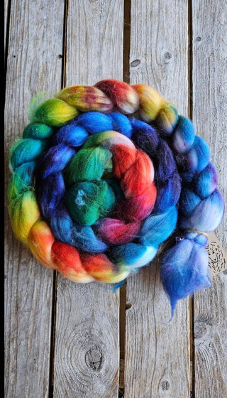 The Koigu Merino Roving Braid, featuring vibrant shades of blue, red, green, yellow, and purple, is displayed in a spiral on a rustic wooden surface.
