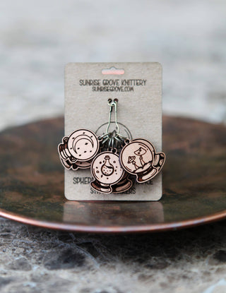 The 🔮 Crystal Ball Stitch Marker Set 🔮 includes three cherry wood markers with mystical designs, displayed on a Sunrise Grove Knittery card—the perfect witchy accessory for your knitting. The card sits on a textured surface with a blurred background.