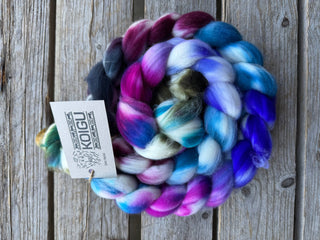 A Koigu Merino Roving Braid, featuring coiled multicolored merino wool in pink, blue, purple, white, and green, rests on a weathered wooden surface with a Koigu tag attached.