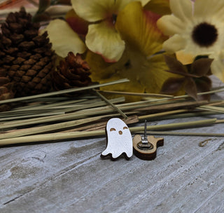 A pair of small wooden ghost earrings rests on a rustic wooden surface among pinecones, dried flowers, and hay-like stems—perfect for fans of handmade jewelry, with sturdy and hypoallergenic earring hooks visible beside them.