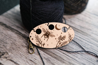 The Yarn Management Tool for Knitters is an oval wooden keychain featuring a charming design of a bee, plants, stars, and a crescent moon, displayed on a wooden surface beside dark yarn balls—making it an ideal crochet accessory or practical knitting tool.