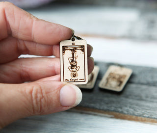 A hand holds a small wooden charm engraved with a tarot card, perfect as a Tarot Card Stitch Marker—choose from The Fool, Death, or Temperance—while two more charms blur in the background on a rustic surface.