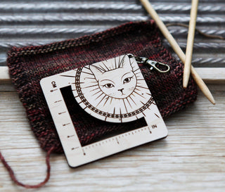 The Witchy Cat Gauge Swatch Ruler, featuring a cat face design, sits on a knitted swatch next to two wooden knitting needles on a wooden surface.