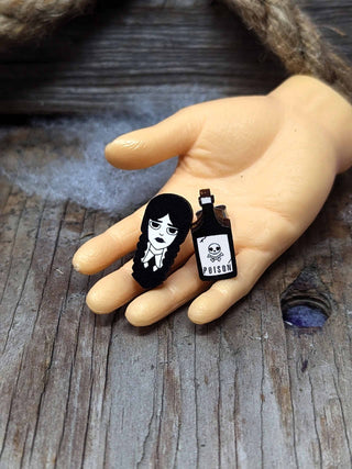 A small plastic hand on wood displays Wednesday Stud Earrings: one features a girl with long black braids and a stern look, the other is a bottle labeled POISON—unique pieces among handmade San Francisco treasures.