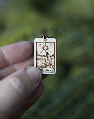 A hand holds a small wooden Tarot Card Stitch Marker featuring The Star, part of the "The Moon, The Star & The Sun" set—an enchanting progress keeper for knitting, with blurred greenery highlighting its charm.