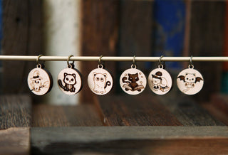 The 🐾 Scaredy Cats Stitch Marker Set 🐾 includes six round maple wood stitch markers with cute Halloween animal designs like a fox witch, skeleton panda, bookish cat, and pumpkin mouse—perfect spooky knitting accessories.