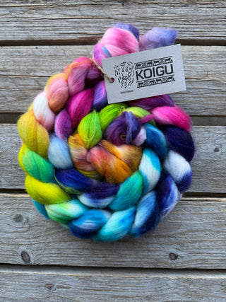 The Koigu Merino Roving Braid, a soft bundle of multicolored merino wool in vibrant pink, green, blue, yellow, and purple shades, rests on a wooden surface with its Koigu tag attached.