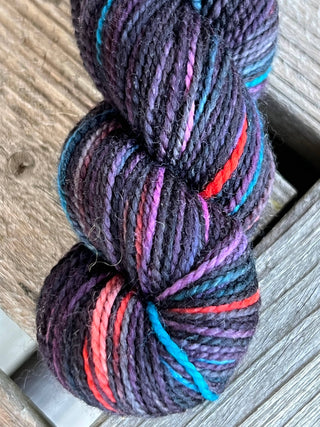 A close-up of a KPM and KPPPM Variegated Color Collection Skein with a dark base, showcasing vibrant streaks of red, blue, purple, and pink on weathered wooden boards—ideal for hand knitting projects.