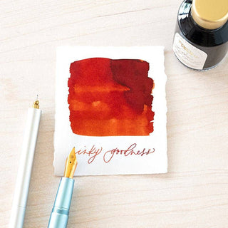 A Bottled Ink and fountain pen rest beside a torn-edged paper on a light wooden surface, featuring a red ink swatch and the handwritten phrase “inky goodness,” showcasing the unique appeal of Bottled Ink.