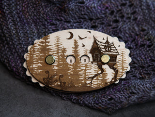 Introducing the Baba Yaga Row Counter: an oval wooden pin engraved with a forest and a house on chicken legs. This wooden knitting row counter features three holes with gold pins and is pictured resting on textured purple knitted fabric.