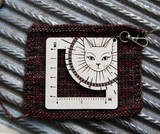 The Witchy Cat Gauge Swatch Ruler features a maple wood cat face with a sunburst design, displays both inch and centimeter markings, and rests on a knitted swatch with gleaming metal ridges in the background.