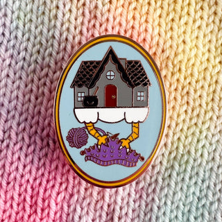 The Baba Yaga Knitting Enamel Pin features a whimsical house on chicken legs, perched on purple knitting with needles and yarn, set against a pastel knitted backdrop.