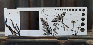 The Luna Moth “Almost Everything” Ruler is a wooden yarn weight tool with rectangular cutout, circular holes, and engraved butterfly, crescent moon, stars, plants, and mushrooms—ideal for fiber arts enthusiasts.