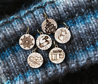The Maple Wood Yuletide Knitting Stitch Markers Set of 6, featuring festive Christmas designs, is displayed on blue and gray knitted fabric.