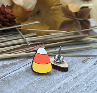 Candy Corn Earrings are displayed on a light wooden surface with autumn decorations. One earring stands upright while the other reveals its metal backing, highlighting their festive Halloween design.