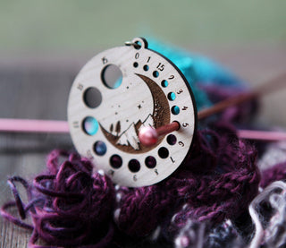 The Crescent Moon Knitting Needle Gauge, featuring crescent moon and mountain etchings, sits on purple and blue yarn as a knitting needle threads through its center; the softly blurred background highlights its design.