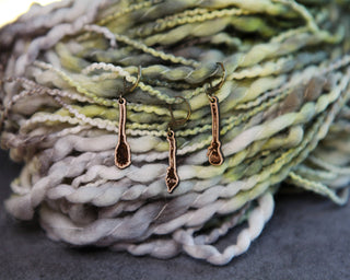 Three small 🧹 Witch’s Broom Stitch Markers 🧹 hang before thick, multicolored yarn in green, gray, and white tones, creating a textured, cozy setting ideal for knitting or crochet projects.