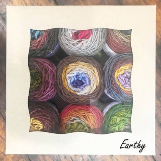 The Freia Minikin Palette Pack in Earthy displays nine colorful merino fingering yarn cakes through a wavy-edged window, perfect for Fair Isle sweaters. "Earthy" is written in cursive on the box’s lower right corner.