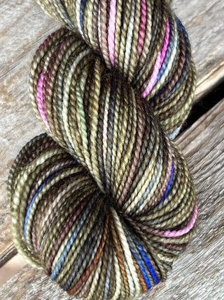 A close-up of a KPM and KPPPM Variegated Color Collection Skein in olive green, brown, pink, blue, and cream hues, twisted and resting on a wooden surface—ideal for hand knitting projects.