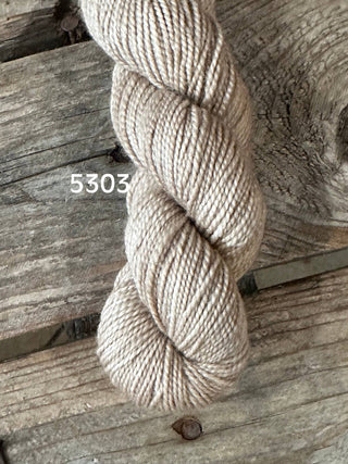 A skein of Koigu KPM & KPPPM Fingering Weight Yarn in light beige, color 5303, rests on a rustic wooden surface with distinct grain and knots.