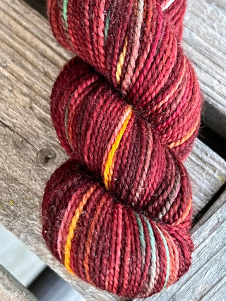 A close-up of a KPM and KPPPM Variegated Color Collection Skein in rich red, orange, yellow, green, and brown hues—perfect for hand knitting—resting on a weathered wooden surface.