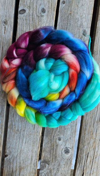 A Koigu Merino Roving Braid in the color "blue rainbow 131"—a coiled bundle of multicolored wool—rests on a weathered wooden surface.