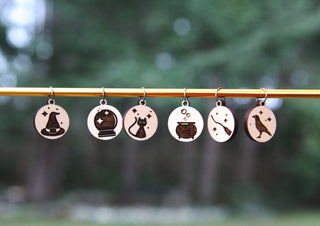 The Witching Hour Stitch Markers set features six round maple wood markers engraved with a witch hat, crystal ball, cat, cauldron, wand, and raven—perfect for adding magical charm to your knitting accessories. Blurred forest background.