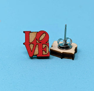 The ❤️ LOVE Earrings ❤️ feature bold red wooden letters standing upright by their backing on a light blue surface—these handmade, hypoallergenic earrings add a charming statement to any outfit.