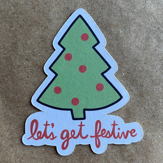 The Let's get festive holiday tree sticker features a green Christmas tree with red ornaments and black outlines, "let's get festive" in red script below, on a brown textured background—printed on eco-friendly paper.