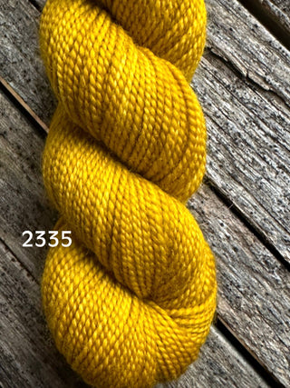 A skein of Koigu KPM & KPPPM Fingering Weight Yarn in bright golden yellow (color 2335) rests on a rustic wood surface, ideal for your next knitting or crochet project.