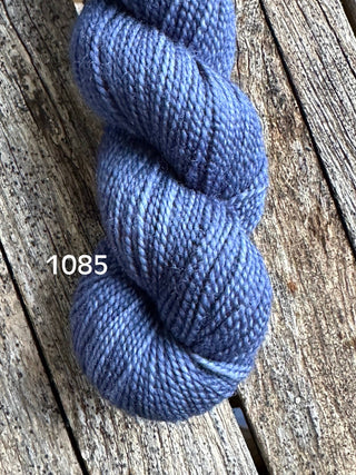 A skein of Koigu KPM & KPPPM Fingering Weight Yarn in blue-purple, labeled 1085, rests on weathered wooden boards, highlighting its soft texture and subtle color shifts—ideal for knitting or crochet projects.