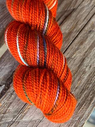 A close-up of a KPM and KPPPM Variegated Color Collection skein in orange with multicolored streaks, resting on weathered wood—ideal for hand knitting projects.