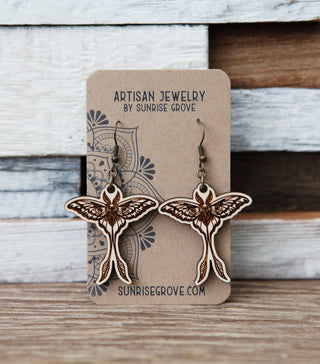 A pair of Luna Moth Maple Wood Earrings with intricate patterns, displayed on a branded card reading Artisan Jewelry by Sunrise Grove, set against a rustic wooden background.