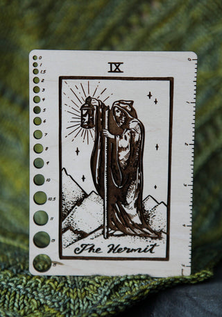✨ The Hermit ✨ Tarot Knitting Needle Gauge & Ruler is a maple wood measuring tool engraved with The Hermit tarot design—hooded figure, lantern, staff, and mountains. Includes ruler markings and circular needle gauge holes along the side.