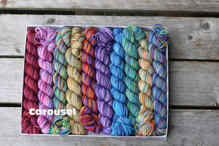The Koigu Pencil Box holds mini skeins of multicolored merino wool yarn in rainbow hues. Carousel is printed in white on the lower left skeins. The box rests on a weathered wooden surface.