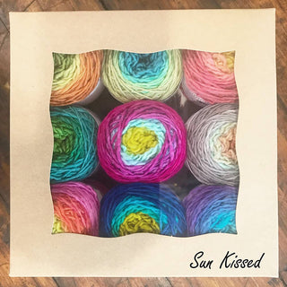 A square box with a wavy-edged window shows nine Freia Minikin Palette Packs yarn cakes in Sun-Kissed, arranged in a grid. The colorful, multicolored yarns are ideal for baby blankets or Fair Isle sweaters. "Sun-Kissed" appears in the bottom right corner.