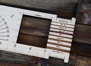 The Butterfly "Almost Everything" Ruler, a wooden tool for knitters and crocheters, displays yarn measured against engraved wraps-per-inch and weight markings—an essential accessory for every maker.