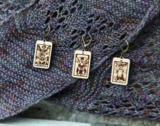 The 🔮 Tarot Card Stitch Markers– The Tower, The Devil & Judgement 🔮 are intricately designed maple wood earrings that double as unique progress keepers or tarot stitch markers, shown on textured purple knit fabric.